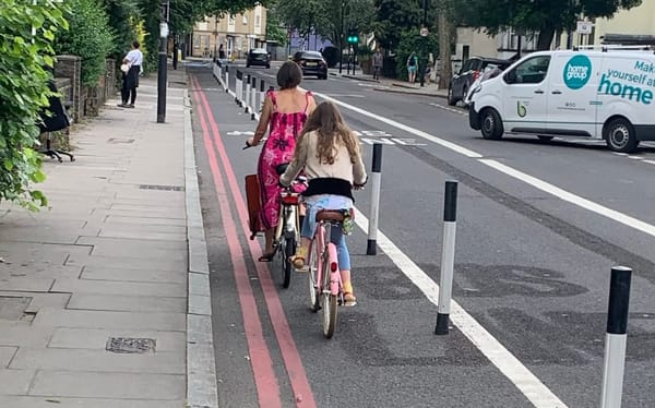 A woman in a dress rides a boxbike in a cycle track protected by platic wands. A teenage girl cycles behind her.